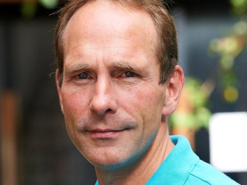 Fotoet viser Peter Viggo Jakobsen, som ser direkte ind i kameraet iført en turkis poloshirt foran en sløret baggrund