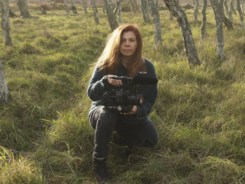 Billedet forestiller en kvinde siddende i græsset mellem unge træer.