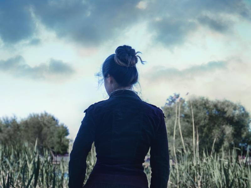 Billedet forestiller en kvinde set fra ryggen. Hun er iført en gammeldags mørk kjole og har sit mørke hår sat op i en knold.
